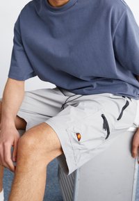 Hombre sentado en una caja gris, vistiendo una camiseta azul y pantalones cortos gris claro con bolsillos con cremallera negros y un logotipo rojo-naranja.