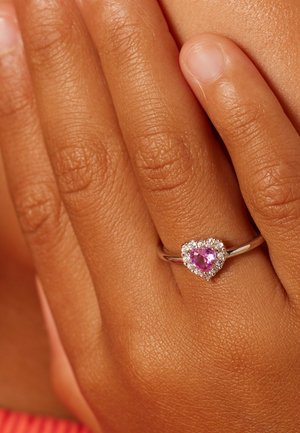 Hand mit mittelbrauner Haut, die an dem Ringfinger einen silbernen Ring trägt, der einen herzförmigen pinkfarbenen Edelstein zeigt, umgeben von kleinen klaren Steinen.