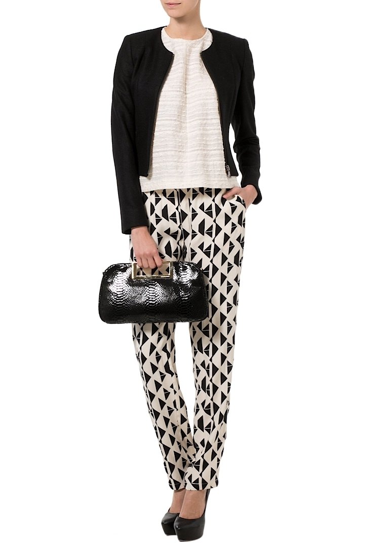 Femme portant une veste courte noire, un top blanc texturé, un pantalon blanc avec des formes géométriques noires, des talons noirs et tenant un sac à main noir texturé.