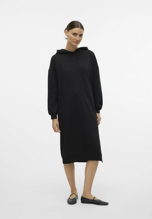 Robe sweat à capuche noire avec des manches longues, coupe décontractée et fentes sur les côtés, associée à des chaussures plates noires. Tissu texturé simple.