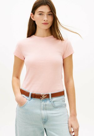 Ung kvinde med langt brunt hår iført en lyserød tætsiddende t-shirt, der er stoppet ned i lyseblå jeans med et brunt bælte med en guldlås.