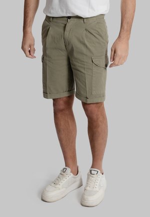 Pantaloni cargo verde oliva con orli risvoltati, diverse tasche laterali, realizzati in tessuto leggero. Indossati con scarpe sportive bianche.