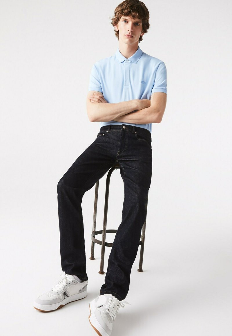 Junger Mann, der auf einem hohen Metallhocker sitzt, trägt ein hellblaues Poloshirt, dunkle Jeans und weiße Turnschuhe vor einem weißen Hintergrund.