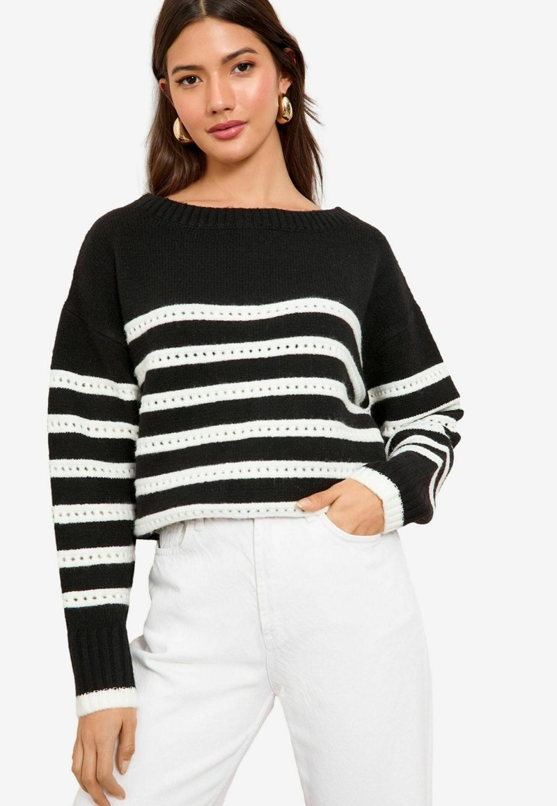 Maglione a righe bianche e nere con scollatura ampia e polsini a costine, caratterizzato da righe orizzontali e dettagli a occhiello. Abbinato a pantaloni bianchi.