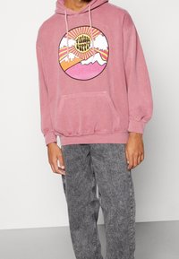 Sudadera rosa con un gráfico circular que presenta montañas y el texto "Vintage Supply". Material de algodón, bolsillo tipo canguro, puños acanalados.