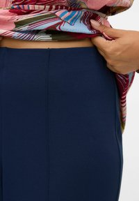 Pantalons bleu marine en tissu lisse, présentant une coupe ajustée avec un détail de couture sur le côté, partiellement cachés par un haut à motifs.