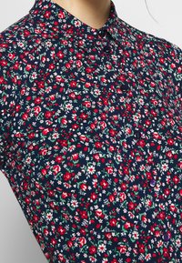 Blommönstrad skjorta tillverkad av lättviktsmaterial, med en marinblå botten och röda samt vita blommor, en spetskrage och knappar.