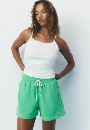 Woman with curly hair wearing a white tank top and green drawstring shorts standing with hands on her hips against a white background.