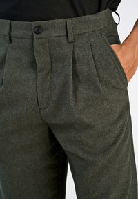 Ciemnozielone spodnie z mieszanki wełny o dopasowanym kroju, z plisami z przodu, szlufkami na pasek i kieszeniami bocznymi. Zblizenie na teksturowany materiał.