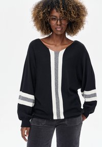 Zwarte gebreide trui met een V-hals, voorzien van witte en grijze verticale strepen en brede mouwen. Gecombineerd met donkergrijze ribfluwelen broek.