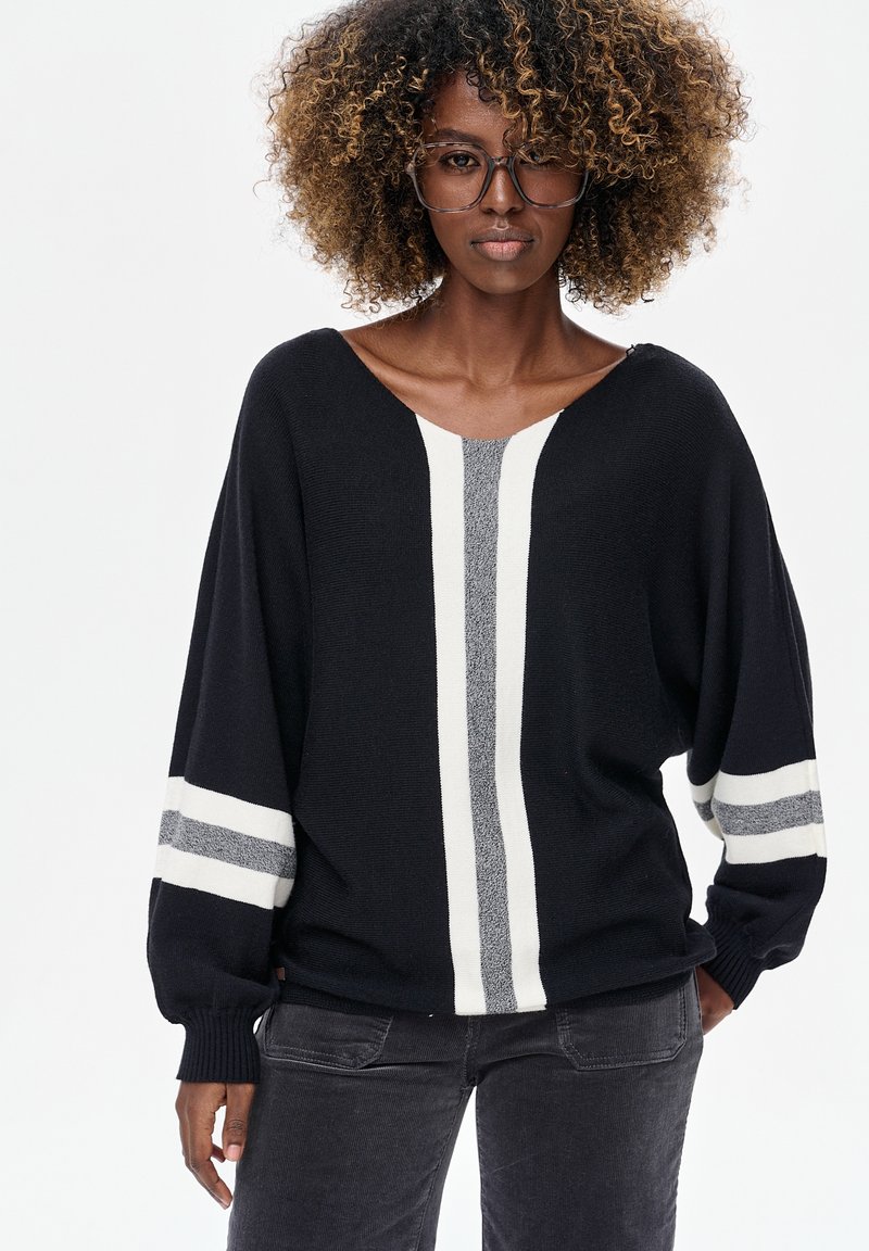 Zwarte gebreide trui met een V-hals, voorzien van witte en grijze verticale strepen en brede mouwen. Gecombineerd met donkergrijze ribfluwelen broek.