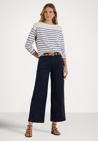 Garās piedurknes svītrains tops krēmkrāsā un zilā tonī, kombinēts ar tumši ziliem, platām biksēm. Apģērbu papildina brūna pītā josta un sandales ar siksniņām.