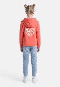 Παιδί με ξανθά μαλλιά σε διπλούς κότσους, φοράει κοραλί φούτερ με κουκούλα με σχέδιο καρδιάς "Peace Amour" στην πλάτη, ανοιχτό μπλε τζιν και λευκά αθλητικά παπούτσια.