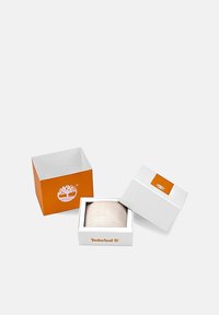 Valge kingituse karp oranžide aktsentidega, millel on Timberlandi logo, sisaldab tekstuuriga beeži eset. Karp on avatud ja näitab sees olevat eset.