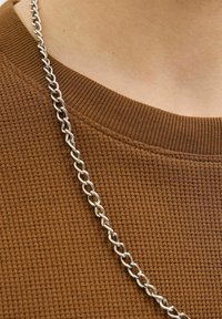 Collar de cadena de metal plateado sobre un suéter marrón texturizado de punto con un cuello acanalado. Se pueden ver los eslabones de la cadena entrelazados.