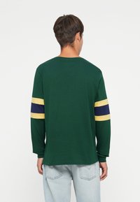 Long-sleeve T-shirt in dark green cotton, featuring yellow and navy striped accents on the sleeves, with a relaxed fit and round neckline.