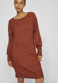 Vestito in maglia di colore arancione bruciato, con maniche lunghe e una vestibilità comoda. Il tessuto ha una consistenza morbida con una leggera lucentezza.
