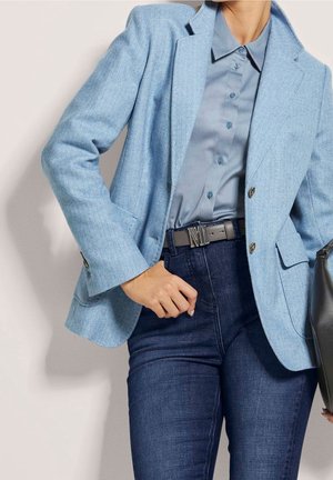 Vrouw die een lichtblauw colbert draagt, een blauw overhemd met knopen, donkerblauwe spijkerbroek en een zwarte riem met zilverkleurige gesp, en een zwarte handtas vasthoudt.