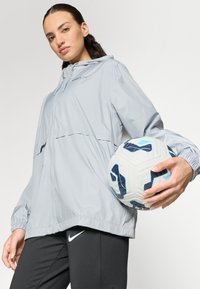 Veste coupe-vent gris clair avec capuche, poignets resserrés et fermeture éclair, assortie à un pantalon de sport noir et un ballon de football blanc.