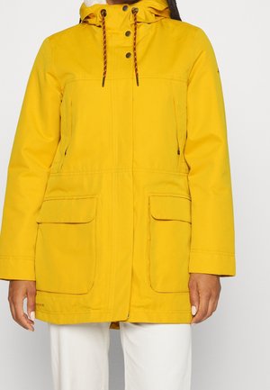 Femme portant une veste à capuche jaune avec poches à rabat à l'avant et boutons noirs, associée à un pantalon blanc, debout devant un fond uni.