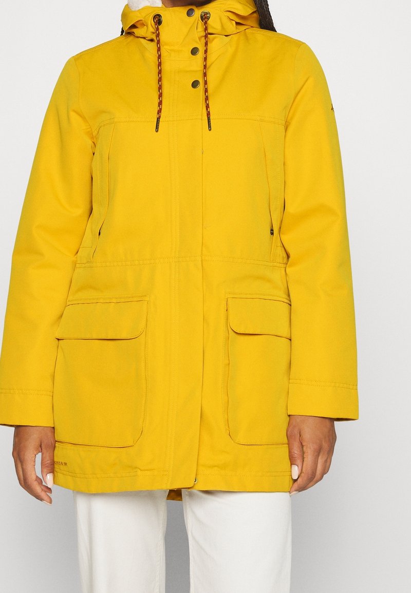 Femme portant une veste à capuche jaune avec poches à rabat à l'avant et boutons noirs, associée à un pantalon blanc, debout devant un fond uni.
