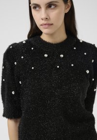 Čierny chlpato sweater s krátkymi rukávmi, okrúhlym výstrihom a roztrúsenými perlovými akcentmi na prednej strane pre textúru a detail.