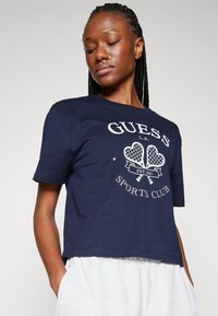 Navy sininen puuvillapaita lyhyillä hihoilla, jossa on valkoinen graafinen printti sydämistä ja mailoista, nimellä "GUESS SPORTS CLUB."