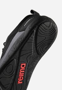 Semelle de chaussure de sport noire avec motif de bande de roulement détaillé et logo "reima" rouge en bas, vue latérale sur fond blanc.
