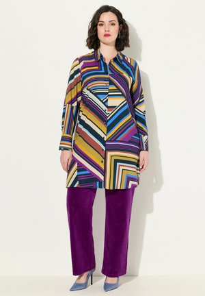 Blouse longue à rayures multicolores avec fermeture à boutons, associée à un pantalon en velours côtelé violet et des talons bleu clair. Texture du tissu lisse.