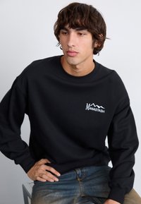 Black sweatshirt made of soft fabric, featuring a white logo with mountain graphics. Relaxed fit with cuffed sleeves, simple and modern design.