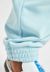 Light blue jogger pants with ribbed cuffs and a smooth texture. Features a close-up view of the cuff and a white sneaker with blue accents.