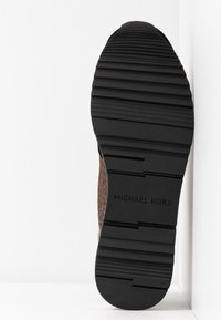 Černá gumová podešev s texturovanými drážkami, na kterých se nachází embosovaná etiketa "MICHAEL KORS" a kontrastní hnědý tkaný detail na vršku.