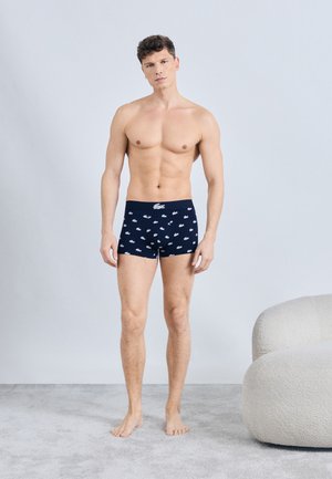 Muškarac bez majice stoji bos na tepihu, noseći mornarskoplave bokserice s bijelim logotipima aligatora, pored zakrivljene bijele fotelje.