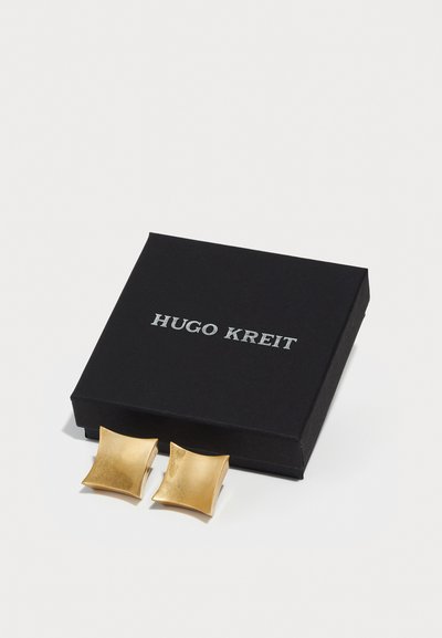 Kuldseid mansettnuppe, millel on poleeritud, laineline ruudukujuline disain, puhkevad musta karbi kõrvale, millel on hõbedaste tähtedega silt "Hugo Kreit".