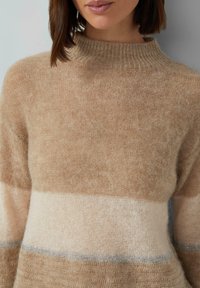 Kobieta nosząca miękki, puszysty sweter w beżowe i kremowe kolory z prążkowanym wysokim dekoltem.