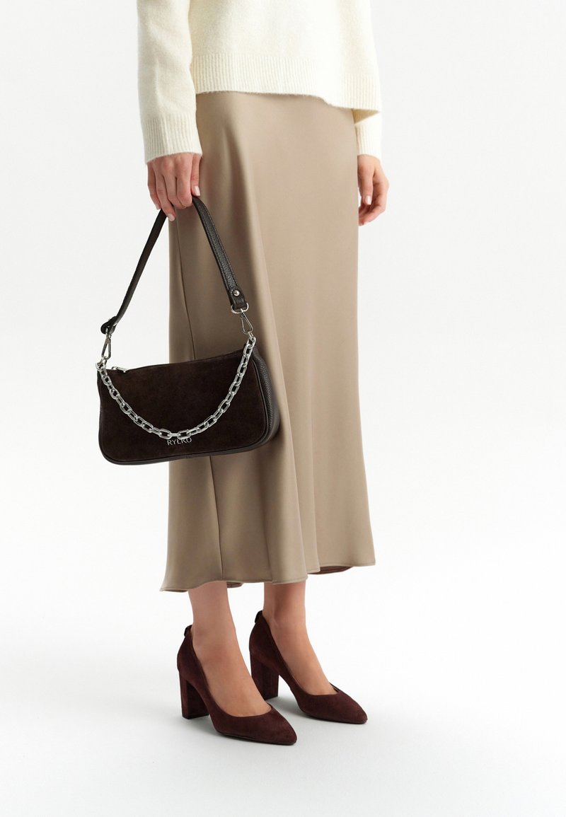 Brązowa torebka z zamszu z srebrnym łańcuchem. Połączona z beżową satynową spódnicą i brązowymi zamszowymi szpilkami na słupku.