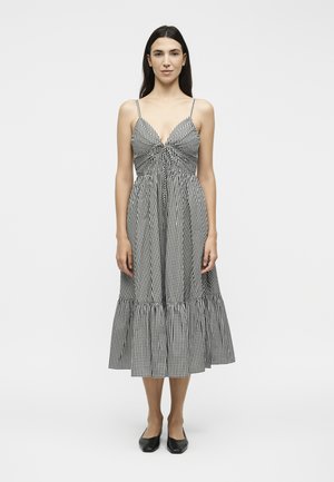 Femme portant une robe midi vichy noire et blanche à fines bretelles et des chaussures plates noires, debout devant un fond clair uni.