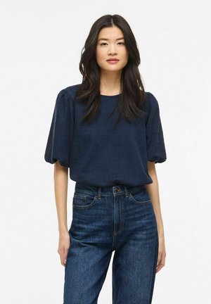 Jeune femme aux longs cheveux bruns portant un chemisier bleu marine à manches bouffantes et un jean bleu taille haute, debout devant un fond blanc.