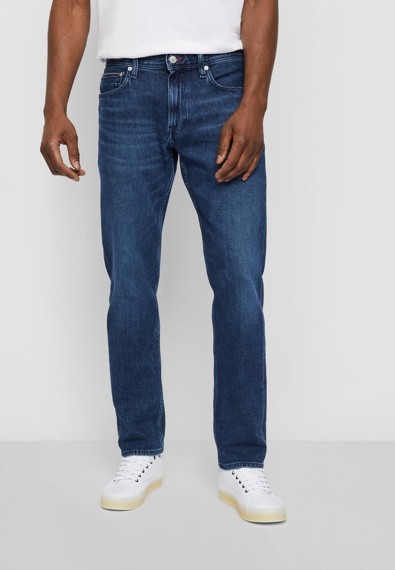 Pantalones vaqueros de mezclilla azul oscuro con un corte recto, que presentan un diseño estándar de cinco bolsillos y un cierre de cremallera. Llevados con zapatillas blancas.