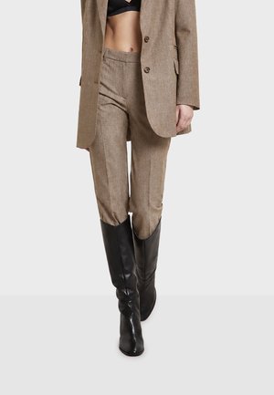 Vrouw die beige op maat gemaakte broek en bijpassend blazer draagt over een zwart cropped top en zwarte kniehoge leren laarzen.