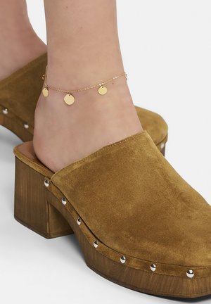 Braune Wildleder-Clogs mit einem klobigen Holzabsatz, die an der Seite mit silbernen Nieten verziert sind. Auch ein goldener Fußkettchen mit Scheiben ist sichtbar.