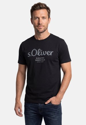Man met kort bruin haar en baard, gekleed in een zwart "s.Oliver Elevated Essentials Since 1989" t-shirt en donkerblauwe spijkerbroek, poserend met één hand in zijn zak.