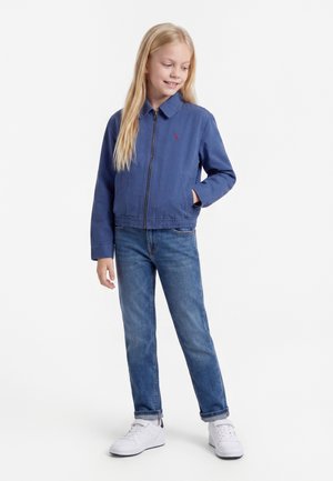 Chica rubia que lleva una chaqueta azul con cremallera, vaqueros azules y zapatillas blancas, de pie con una mano en el bolsillo, sonriendo y mirando de lado.