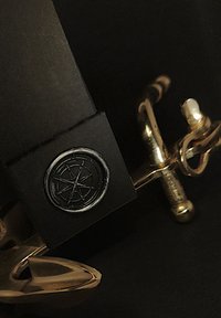 Zwart rechthoekig object met een reliëf cirkelvormig logo met een stervorm, naast een glanzend gouden metalen ankerdetail.