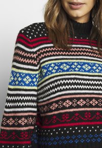 Mångfärgad stickad tröja med svarta, röda, rosa, gula, blå och vita ränder som har geometriska mönster och hög krage.