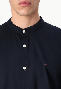 Mand iført en marineblå skjorte med mandarinkrave, hvide knapper og et lille rødt, hvidt og blåt logo på brystet.