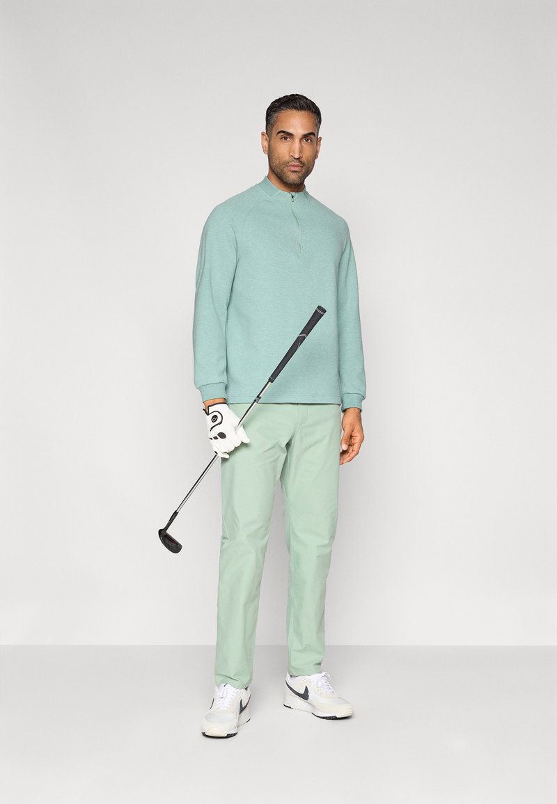 Pull-over vert clair à manches longues avec une demi-fermeture éclair, associé à un pantalon vert clair et des baskets blanches. Tenant un club de golf, portant un gant blanc.