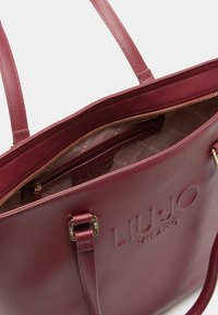 Didelė bordo spalvos rankinė iš tekstūruotos sintetinės medžiagos, su iškilmingu logotipu, užtrauktuko uždarymu ir dviem petiniais diržais. Viduje yra raštuotas pamušalas.