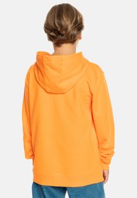 Sudadera con capucha naranja de corte relajado, textura suave y puños acanalados. Presenta un diseño sencillo y una gran capucha en la parte trasera.