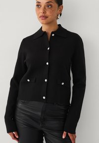 Zwarte gebreide cardigan met een kraag, lange mouwen en zilveren knopen. Twee voorzakken met bijpassende accenten.